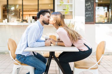 Side view of boyfriend and girlfriend holding holding hands while kissing at sidewalk cafe