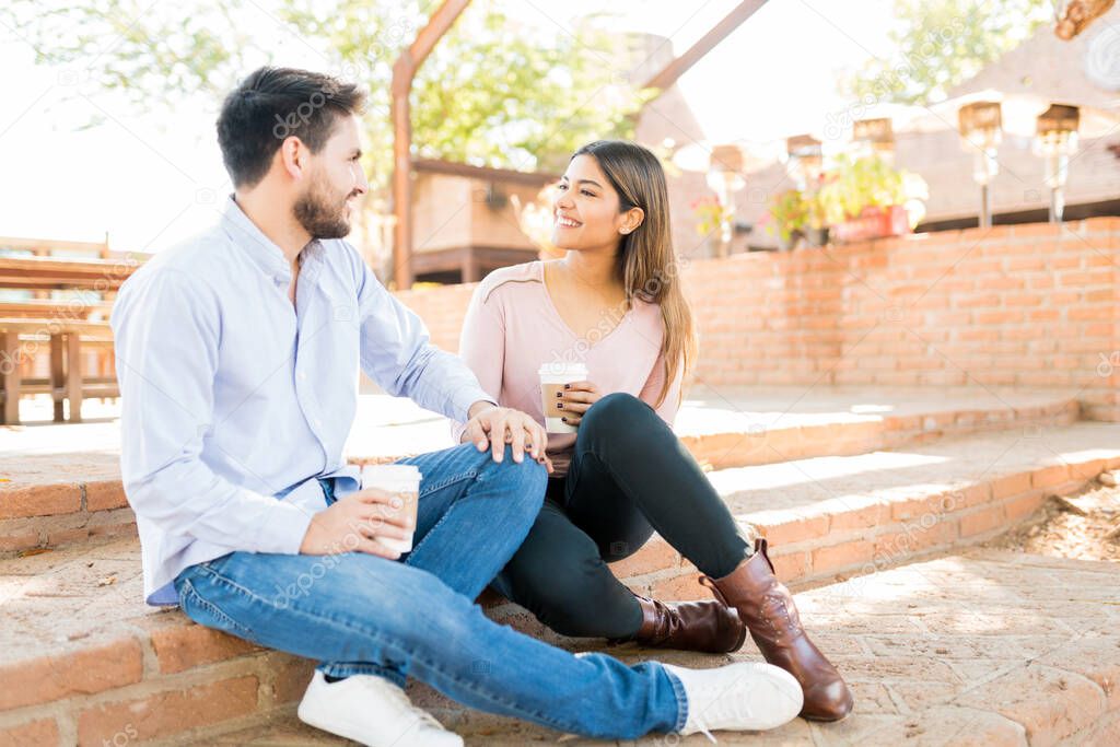 Smiling Latin boyfriend and girlfriend holding disposable coffee cups while sitting on steps outside cafe