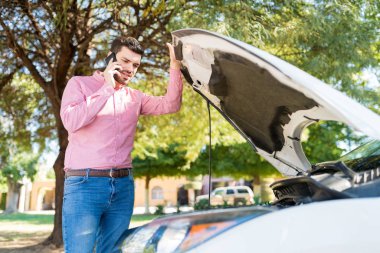 Yetişkin bir adam yol kenarında bozuk bir arabada beklerken cep telefonuyla konuşuyor.