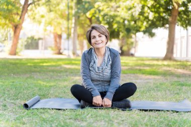 Parktaki yoga minderinde bacak bacak üstüne atmış, gülümseyen emekli yaşlı kadın portresi.