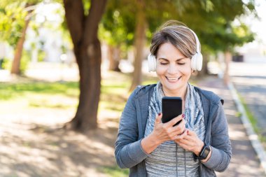 Cheerful active elderly woman looking at mobile phone while listening through headphones on sidewalk