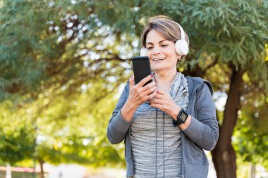 Happy senior woman using smartphone while listening through headphones against trees