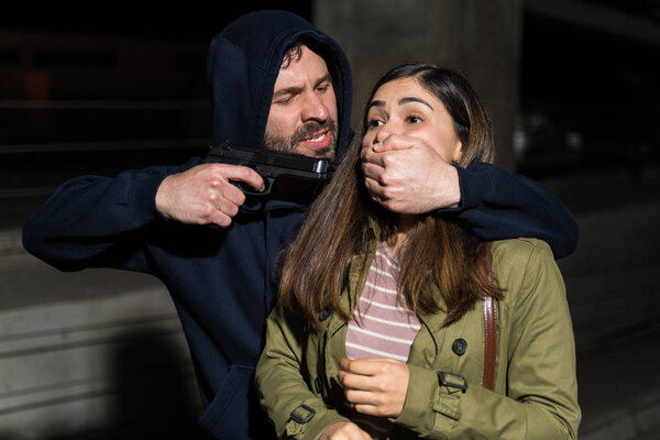 Young woman with mouth covered by criminal holding gun in alley