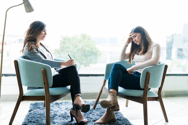 Female counselor writing on notepad while depressed woman sitting with head in hand at office during therapy session