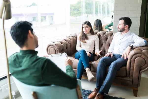 Angry woman sitting with husband while explaining relationship problem to psychologist in office