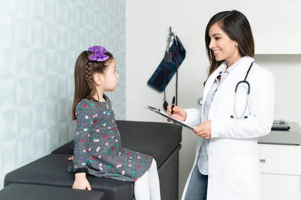 女儿科医生在剪贴板上写医疗记录时与小女孩交谈 — 图库照片