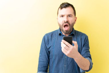 Şok olmuş Latin adam sarı arka planda cep telefonu tutuyor