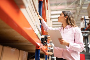 Genç çekici bir çalışan, fabrikada envanter panosunu tutarken rafta inceleme kutusunu tutuyor.