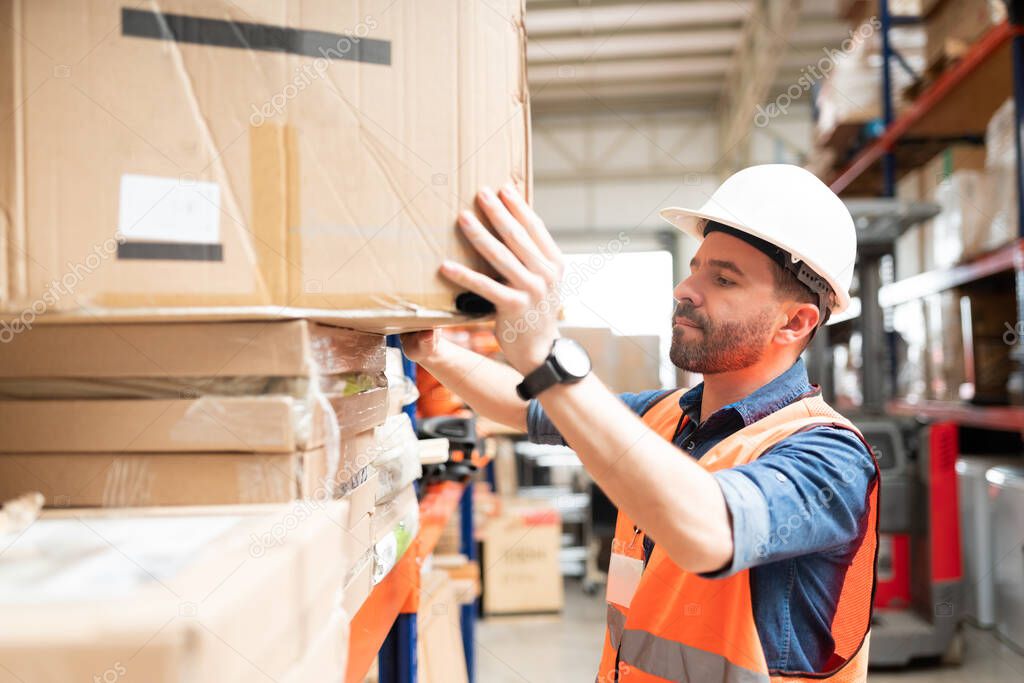 Hispanic worker pushing cardboard box on stack in rack while working at factory