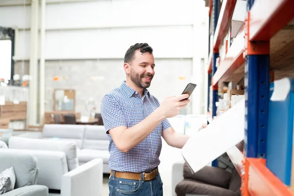 Happy Latin man photographing box in rack through smartphone at warehouse store