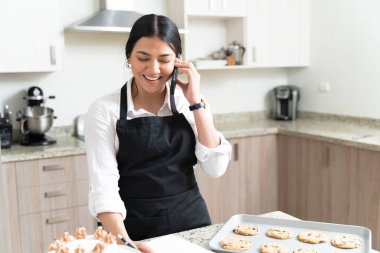 Gülümseyen çekici kadın pasta şefi mutfakta akıllı telefonla konuşurken sipariş yazıyor.