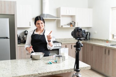 Çekici genç kadın etkileyicisi mutfakta pasta hazırlarken kameradan video bloğu yapıyor.
