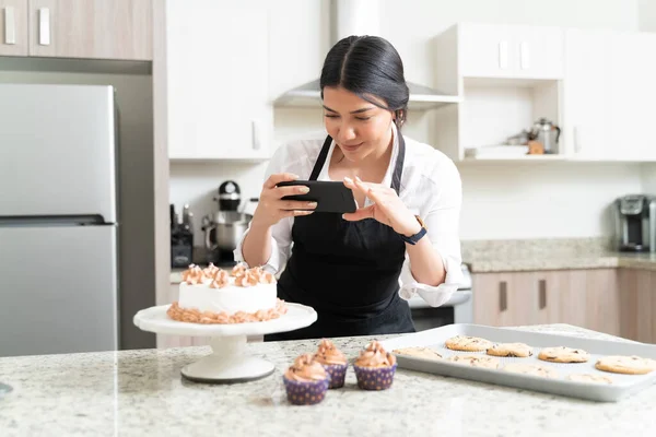 Beautiful female food photographer taking pictures of cake from smartphone for social media in kitchen