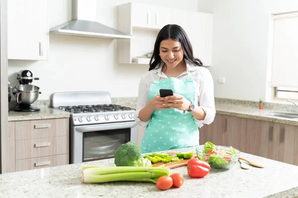 漂亮的家庭主妇在家里厨房柜台边吃蔬菜边用智能手机 — 图库照片