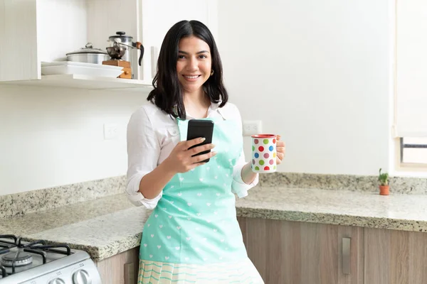 Kahve fincanı ve cep telefonuyla mutfakta duran çekici genç bir kadının portresi.