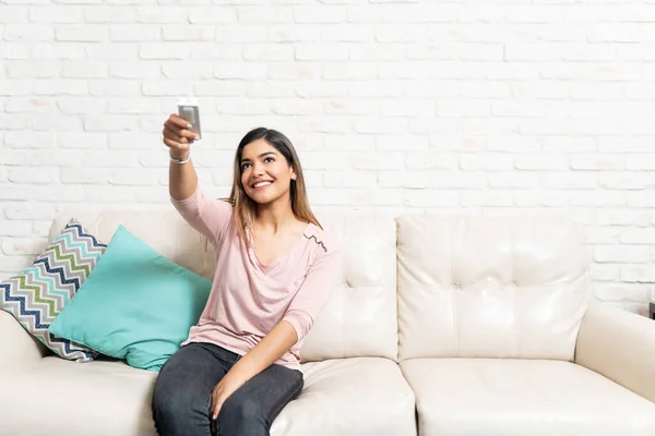 坐在沙发上靠着砖墙 用空调遥控装置微笑美丽的拉丁女人 — 图库照片