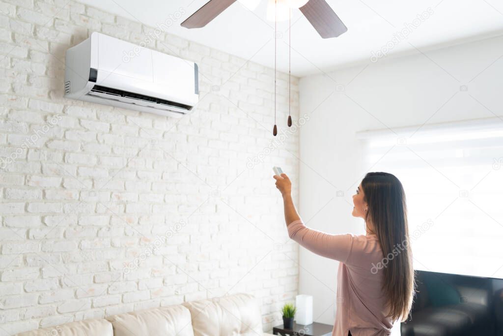 Hispanic young woman adjusting temperature of air conditioner using remote control in room at home