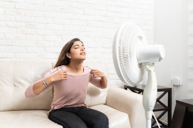 Latin genç bir kadın evdeki sıcak havada kanepede otururken kendini elektrikli vantilatörle serinletiyor.