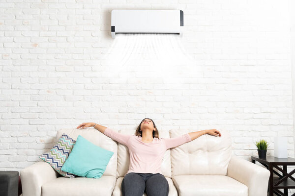 Latin young woman relaxing under mini split on sofa in living room at home