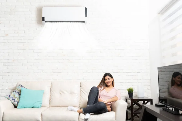 Smiling Hispanic beautiful woman resting on sofa below air conditioner at home