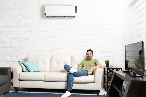 Smiling Hispanic man resting on sofa below air conditioner at home