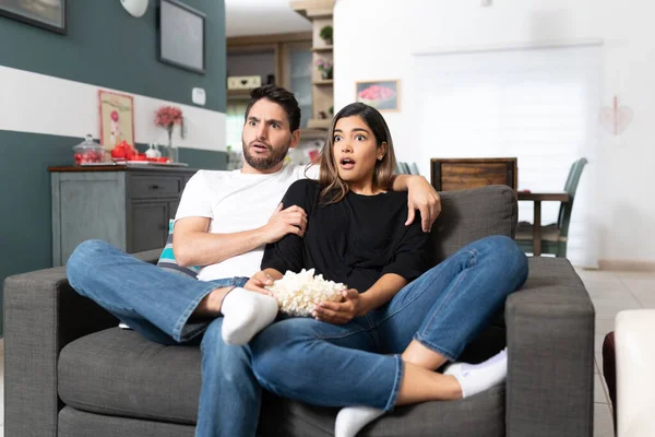 Hispanic couple watching horror movie while sitting on sofa in living room at home