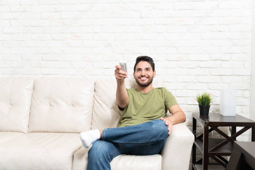 Smiling handsome Latin man using air conditioner remote control while sitting on sofa against brick wall at home