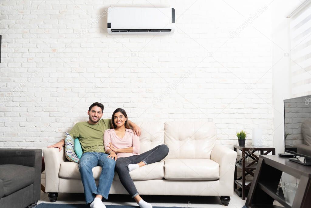 Portrait of smiling Hispanic heterosexual couple relaxing on sofa in living room at home with air conditioning