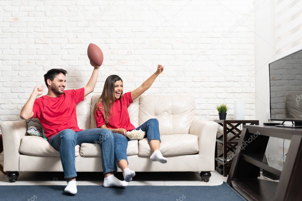 Happy Latin boyfriend and girlfriend watching American football match on TV at home