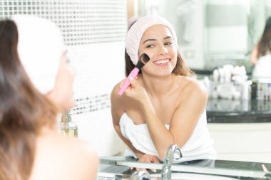 Makyaj fırçası kullanan ve banyo aynasının önünde gülümseyen beyaz bir kadın.