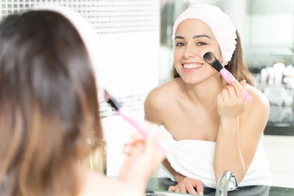 在浴室镜子前使用化妆品刷并进行眼神交流的快乐的白种女人 带着头巾 — 图库照片