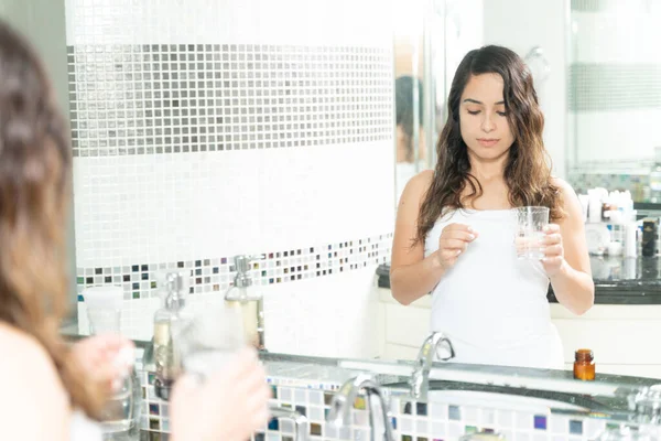 一个漂亮女人在浴室里拿着一颗药丸和一杯水的倒影 — 图库照片