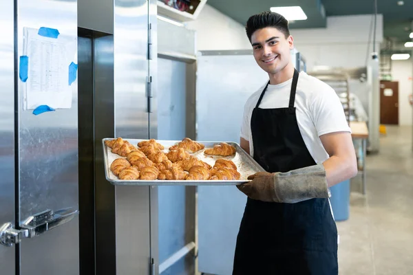 迷人的男性面包师 戴着烤箱手套 手里拿着一个盛满热腾腾的羊角面包的盘子 面带微笑 — 图库照片