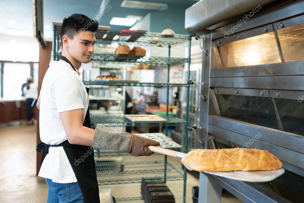 Good looking male Hispanic baker holding a peel with a freshly baked loaf of bread next to the oven