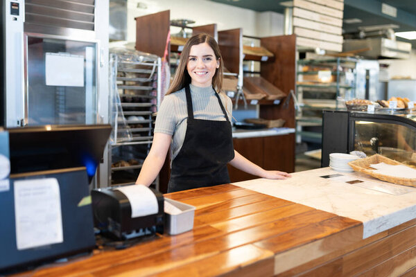 Female baker and entrepreneur standing behind the checkout counter of her bakery shop and smiling