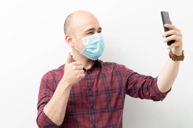Cerrahi maske takan yakışıklı bir adam stüdyoda akıllı telefonuyla video görüşmesi yapıyor.