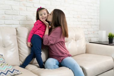 Güzel küçük kız evde dinlenirken annesinin yanağından öpüyor.