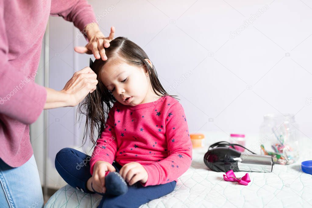 Cute little girl sitting on her bed while her mother combs her hair and works on a hairstyle