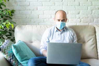 19. koronavirüs salgını sırasında evde dizüstü bilgisayar üzerinde çalışırken ameliyat maskesi takan 30 'lu yaşlarında bir adamın portresi.