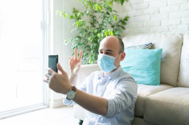 30 'lu yaşlardaki beyaz bir adamın portresi evde cerrahi maske takıyor ve 19 numaralı karantina sırasında akıllı telefonuyla arkadaşlarına merhaba diyor.