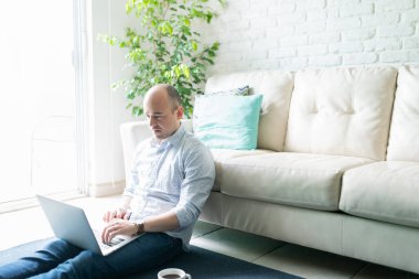 30 'lu yaşlarda yakışıklı bir adam halı döşemesinde oturuyor ve evde dizüstü bilgisayar kullanıyor.