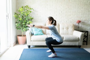 Full length view of an active woman in her 40s working out and doing some squats at home clipart