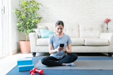Güzel bir esmer kadın evde yoga minderi üzerinde egzersiz yaparken akıllı telefon kullanıyor.