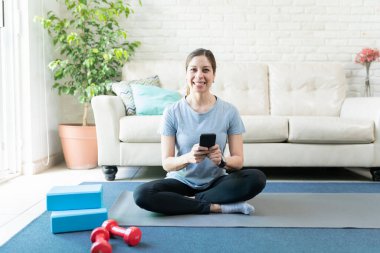 Evde yoga minderinde çalışırken akıllı telefon kullanan gülümseyen aktif bir kadının portresi.