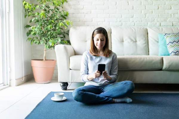 かなりブルネット女性座っていますでザカーペットフロアにホームとチェックソーシャルメディアで彼女のスマートフォン — ストック写真