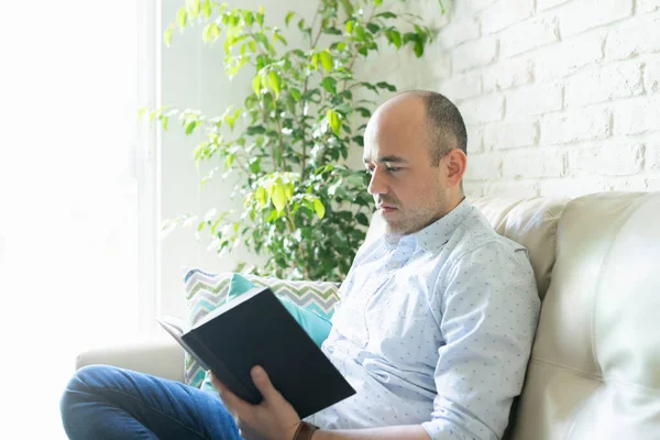 一个30多岁的男人坐在家里的沙发上 穿着休闲装 阅读一本好书的画像 — 图库照片