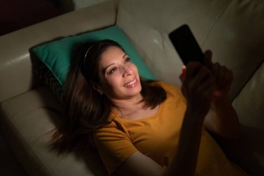 Esmer bir kadının, erkek arkadaşına ya da ortağına gece akıllı telefondan mesaj gönderip gülümsemesini yüksek açılı görmek.