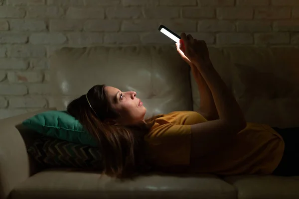 一个白人女人躺在沙发上 晚上看智能手机的照片 — 图库照片