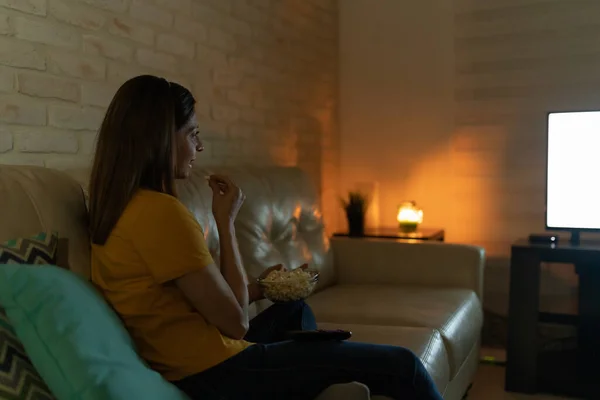 夜にソファに座っている間にブルネットの女性はポップコーンを食べ テレビ番組を楽しむ — ストック写真