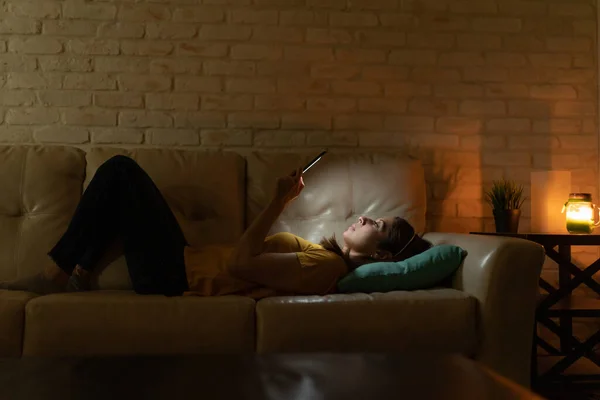 Profile view of a woman lying on a couch and reading a book on an e-reader at night